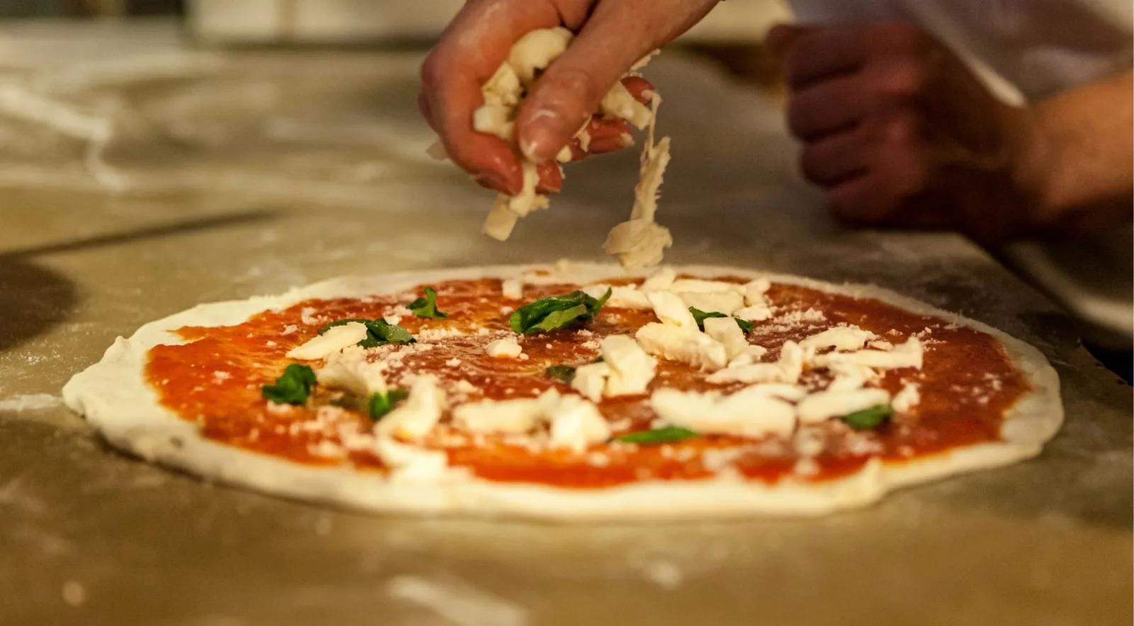 Hands tearing buffalo mozzarella onto a Neapolitan pizza base with San Marzano tomato sauce and fresh basil