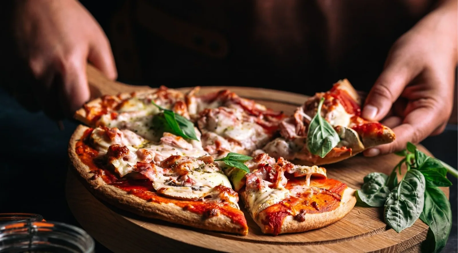 Hands serving sliced pizza topped with prosciutto, mozzarella and fresh basil from a rustic wooden board