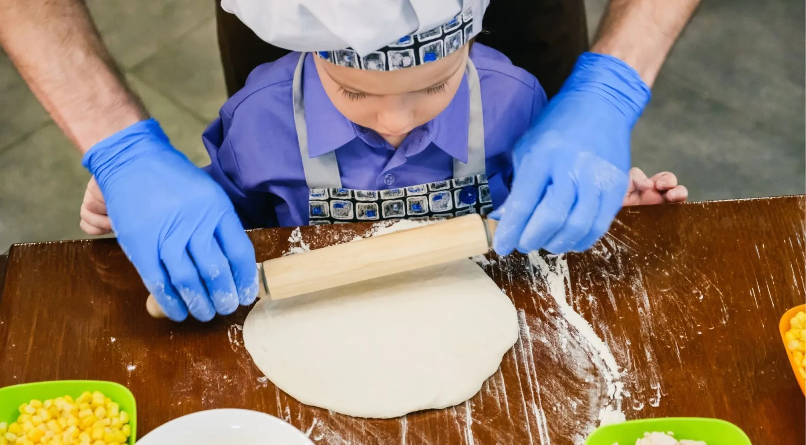 A young child wearing a chef's hat and apron rolling pizza dough with a wooden rolling pin — family-friendly pizza class