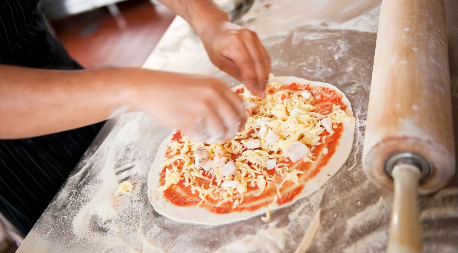 Hands adding tomato sauce and cheese toppings to a pizza base on a flour-dusted surface with a rolling pin beside it