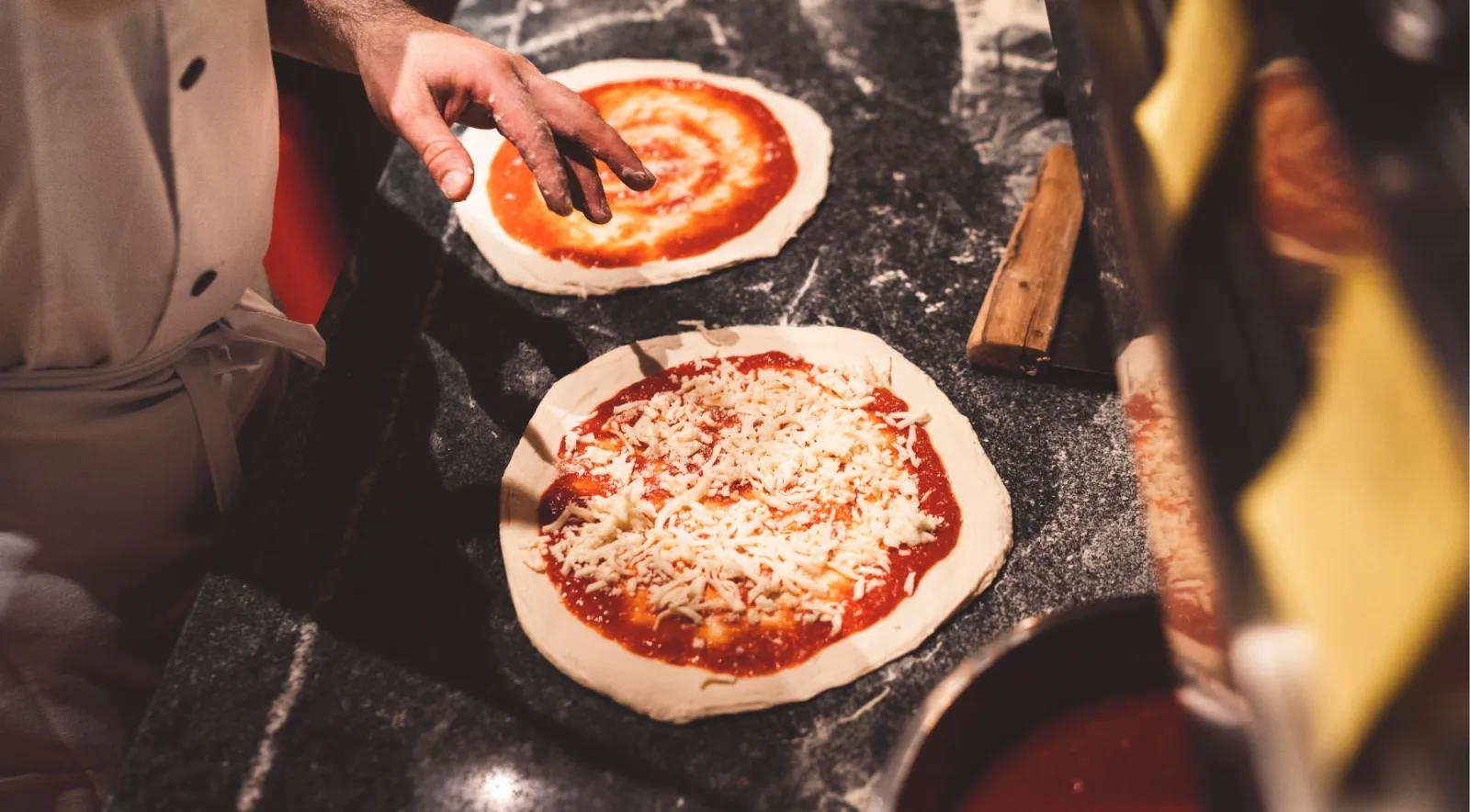 A Neapolitan chef spreading tomato sauce in the traditional circular spiral motion on two pizza bases at a marble counter
