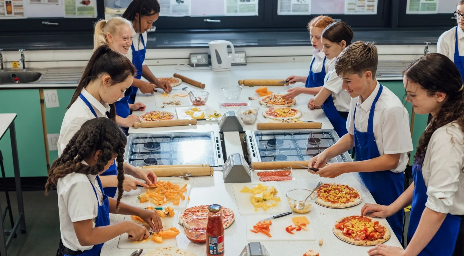 Group of children in blue aprons adding colourful toppings to their own pizzas during a hands-on cooking class