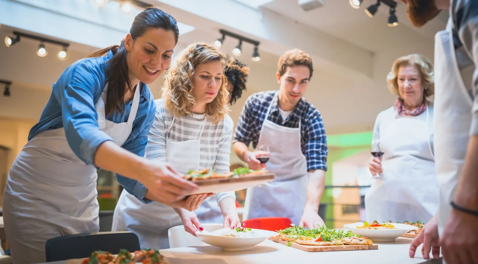 Adults in white aprons working together around a kitchen table during an Italian cooking class with wine glasses in hand