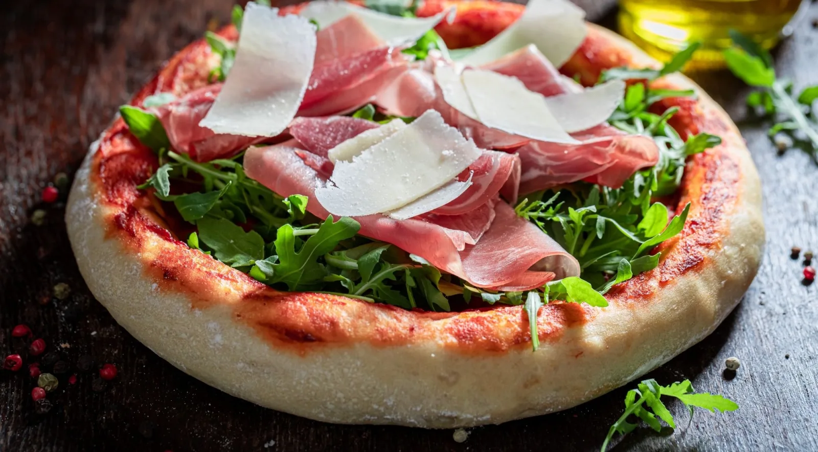 Italian pizza topped with prosciutto crudo, fresh rocket leaves and shaved Parmesan on a rustic dark wooden board