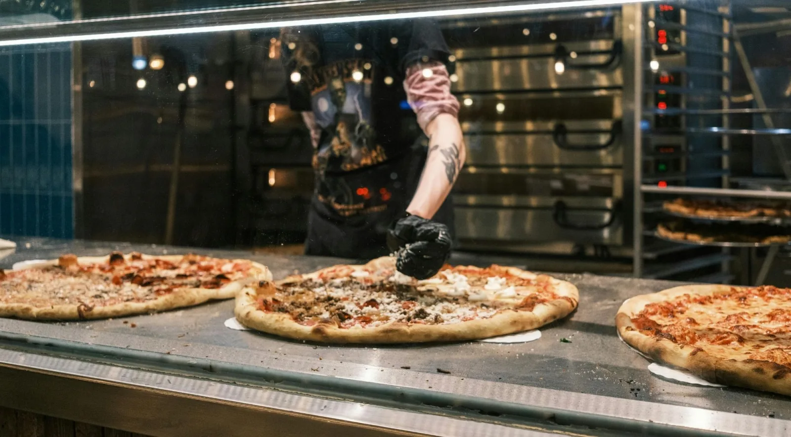 A professional pizzaiolo finishing multiple pizzas at a pizza station with commercial ovens glowing in the background