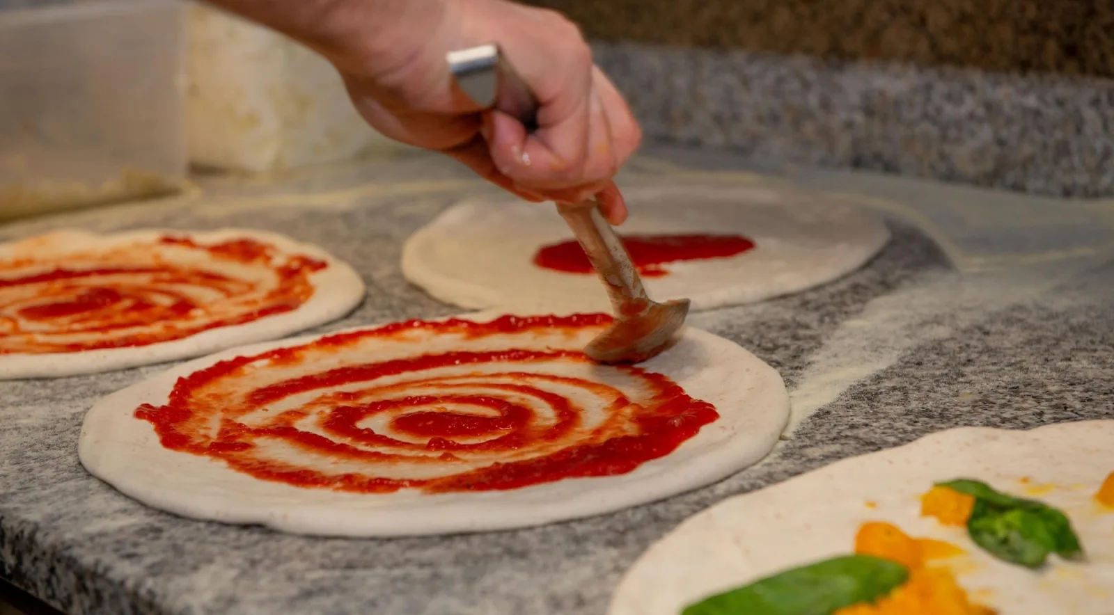 Spreading San Marzano tomato sauce in circular spirals on multiple stretched pizza bases on a marble surface