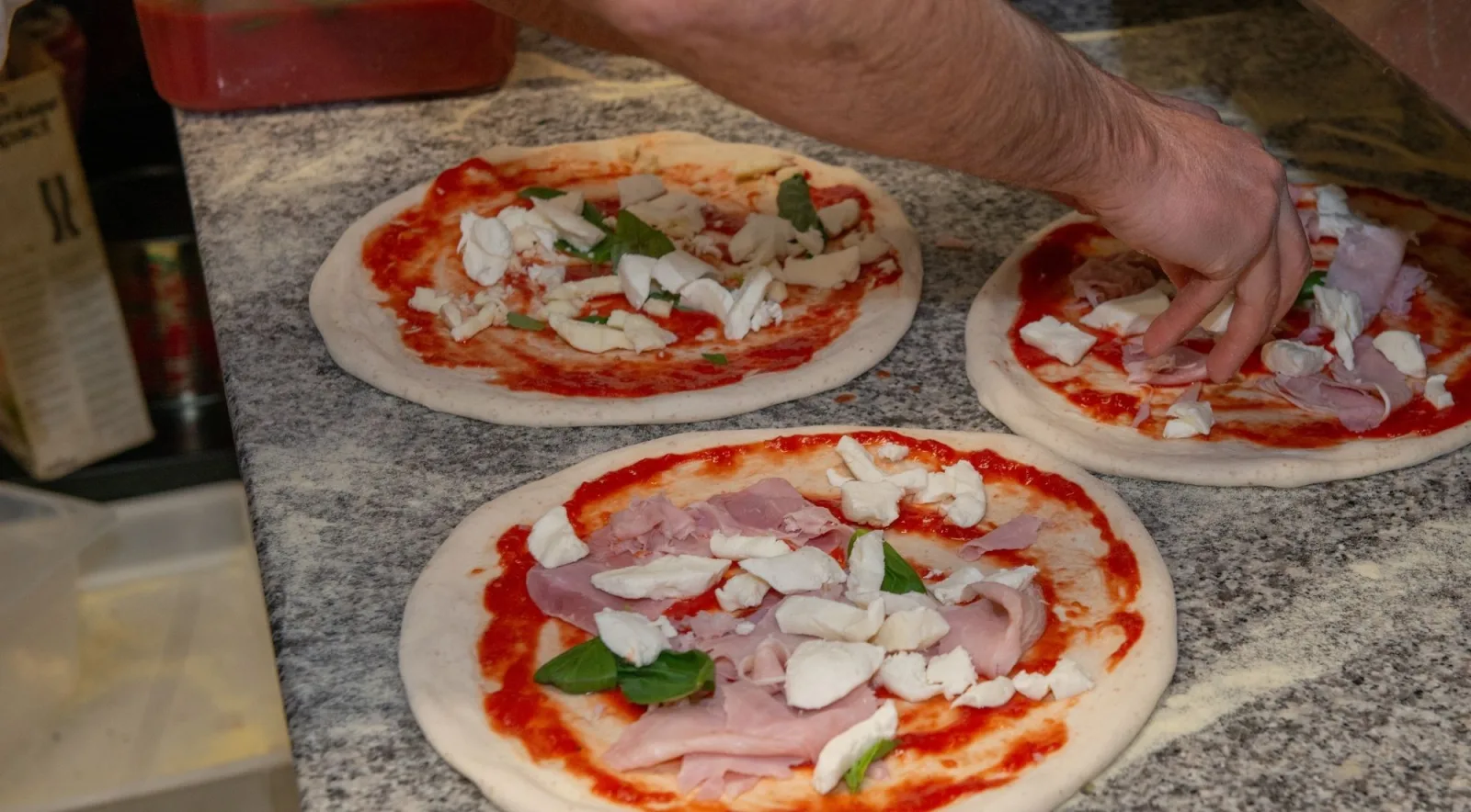 Three pizza bases being topped with tomato sauce, chunks of buffalo mozzarella, prosciutto cotto and fresh basil before baking