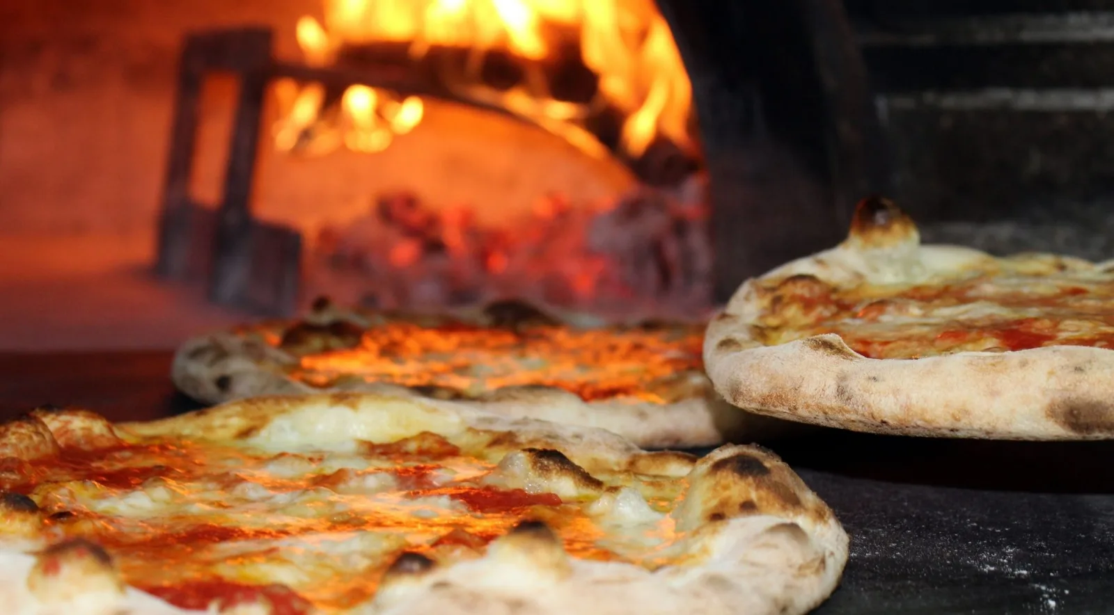 Two Neapolitan pizzas baking on the floor of a traditional wood-fire oven with blazing flames roaring in the background