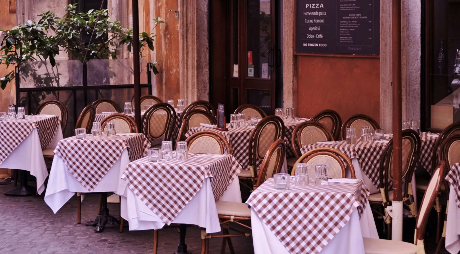 Outdoor terrace of an Italian trattoria with classic checkered tablecloths and a handwritten pizza menu on the wall